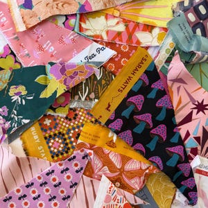 May include: A collection of colorful fabric scraps, many cut into triangular shapes. The fabrics feature various patterns, including floral, mushroom, and geometric designs. Some pieces have text labels, such as "SARAH WATTS" and "RUBY STAR".