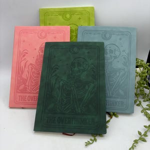 May include: Four journals in various colours, featuring embossed skeleton designs. The journals are in shades of green, pink, blue, and green. The text "THE OVERTHINKER" and "THINKER" are visible. The journals are stacked on top of each other.