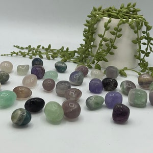 May include: A collection of tumbled fluorite stones in various shades of green, purple, and pink. The stones are smooth and polished, and they are arranged on a white surface.