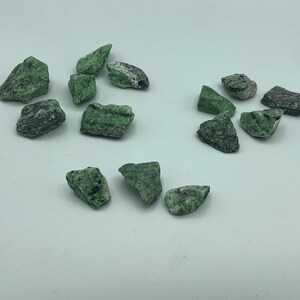 May include: A collection of small, green and black stones with a rough, irregular texture. The stones are scattered on a white surface.