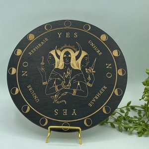 May include: A circular, black wooden divination board with gold etched designs. The board features a central illustration of a three-faced figure and is inscribed with phrases like "YES," "NO," and "UNSURE." Moon phase symbols encircle the edge.