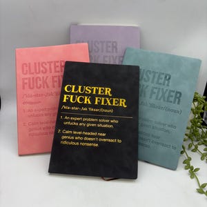 May include: A selection of notebooks in pastel shades of pink, lilac, and teal. Each notebook displays the text "CLUSTER FUCK FIXER". The black notebook at the front has a definition printed on it, with the text in gold.