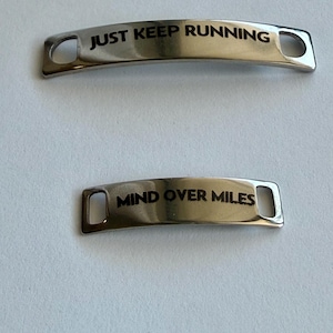 Puede incluir: Dos etiquetas metálicas curvas de color plateado para cordones con acabado pulido. La etiqueta más grande tiene grabado el texto negro "JUST KEEP RUNNING" y la más pequeña "MIND OVER MILES". Ambas etiquetas presentan ranuras rectangulares en los extremos.
