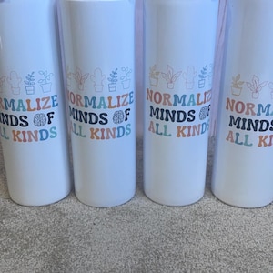 Puede incluir: Vasos aislados blancos con tapas plateadas. Cada vaso presenta el texto "Normalize Minds of All Kinds" en una fuente retro colorida, junto con ilustraciones de plantas. Los vasos están diseñados para mantener las bebidas calientes o frías.