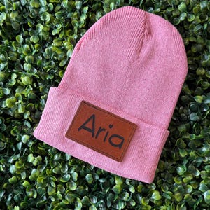 Puede incluir: Gorro de punto rosa con un parche de cuero marrón que dice "Aria" con un pequeño corazón.