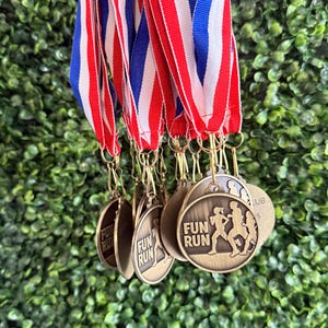 May include: A collection of round bronze-toned medals hanging from red, white, and blue striped ribbons. Each medal features a raised graphic of two people running and the text "FUN RUN" embossed on the front surface of the circular metal disc.
