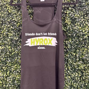 May include: Dark gray racerback tank top with the text "friends don't let friends HYROX alone." in white and lime green. The tank top is hanging on a black hanger against a green textured background.