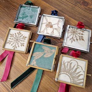 May include: Six square glass ornaments with gold frames and various designs, including snowflakes and botanical patterns. Each ornament has a velvet ribbon in colours like red, green, and blue for hanging. The ornaments are displayed on a wooden surface.