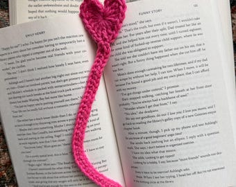 May include: A handmade, hot pink crochet heart bookmark with a long, slender stem, resting on the open pages of a book. The bookmark is a charming accessory for book lovers, adding a touch of handmade flair to reading.