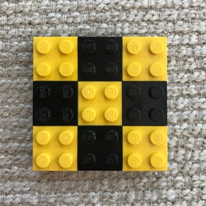 May include: A 3x3 checkerboard made of LEGO bricks. The bricks are yellow and black, alternating colors. The bricks have the LEGO logo printed on them.