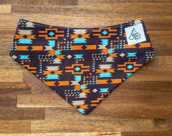 May include: A brown dog bandana featuring a geometric design in shades of orange, teal, and beige. It has a white label with a logo and text. The bandana is made of fabric and is triangular.