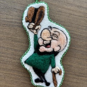 Puede incluir: Adorno de fieltro con un personaje de dibujos animados con chaqueta verde, pantalón negro y sombrero marrón. El personaje sostiene un bastón y tiene una cara blanca con boca roja. El adorno está bordeado con costuras verdes.
