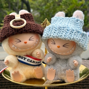 May include: Two plush figures with smiling faces and bunny ear hats. One figure is beige with a brown hat and a "BOY CARTER" banner. The other is gray with a light blue hat. Both are sitting on a gold-colored tray.