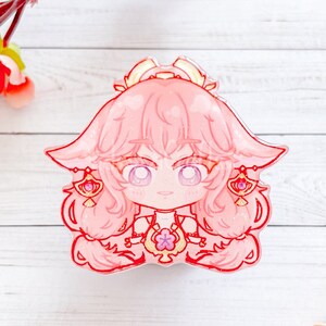 May include: Pink anime-style character with long hair and a flower crown. The character is wearing a white dress with a pink flower design. The background is a white wooden surface.