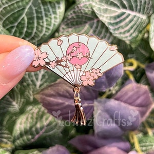 May include: A light blue enamel pin in the shape of a Japanese fan, featuring a pink sun, cherry blossoms, and a tassel. The fan design is detailed with gold accents and black lines.