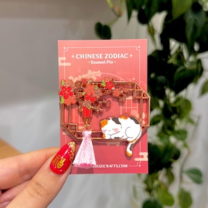 May include: A rose gold enamel pin featuring a sleeping calico cat, a flowering branch, and a pink tassel. The pin is on a pink card with the text "CHINESE ZODIAC - Enamel Pin -". The website address is visible at the bottom.