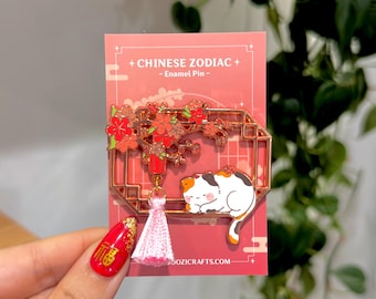 Könnte beinhalten: Ein roségoldener Emaille-Pin mit einer schlafenden Schildpattkatze, einem blühenden Zweig und einer rosa Quaste. Der Pin befindet sich auf einer rosa Karte mit dem Text "CHINESISCHER ZODIAK - Emaille-Pin -". Die Website-Adresse ist unten sichtbar.