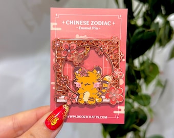 Könnte beinhalten: Eine goldfarbene Emaille-Anstecknadel mit einem Cartoon-Tiger mit rosa Blumen und einem dekorativen Rahmen. Die Anstecknadel trägt die Aufschrift "Chinese Zodiac - Enamel Pin - www.doozicrafts.com".