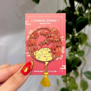 May include: A gold-colored enamel pin featuring a yellow snake within a circular frame adorned with red flowers. The pin is attached to a red card with the text "+ CHINESE ZODIAC + - Enamel Pin -" and a yellow tassel.