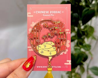 Könnte beinhalten: Ein goldfarbener Emaille-Pin mit einer gelben Schlange in einem kreisförmigen Rahmen, der mit roten Blumen verziert ist. Der Pin ist an einer roten Karte mit dem Text "+ CHINESE ZODIAC + - Enamel Pin -" und einer gelben Quaste befestigt.