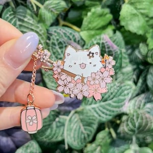 Puede incluir: Un pin de esmalte dorado con un gato blanco con una flor rosa en la cabeza, sentado en un cerezo en flor rosa. El pin tiene un encanto colgante de una linterna rosa con un diseño de flor de cerezo.