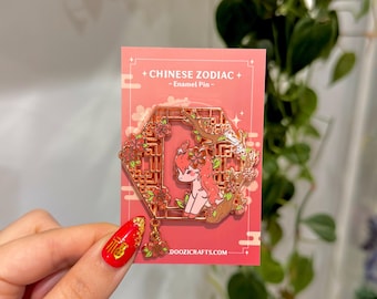 Könnte beinhalten: Eine goldfarbene Emaille-Anstecknadel mit einem Cartoon-Hirsch in einem rosa und goldenen Rahmen mit einem rosa Blumendesign. Die Anstecknadel ist mit "Chinese Zodiac - Enamel Pin - doozicrafts.com" beschriftet.