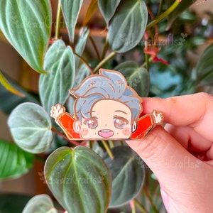 May include: Gold-tone enamel pin featuring a cartoon character with grey hair, wearing a red jacket and smiling. The character has large eyes and a small mouth. The pin is set against a green leafy background.