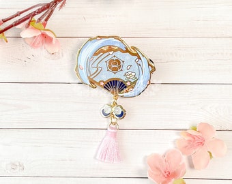 Op de afbeelding: Gouden emaille pin met een blauwe en witte waaier met een bloemenmotief en een roze kwast. De pin is op een witte houten achtergrond met roze bloemen.