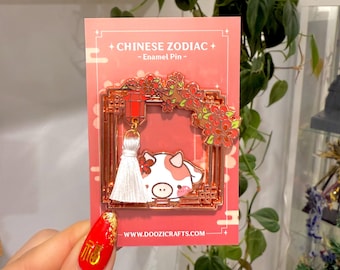 Könnte beinhalten: Eine goldfarbene Emaille-Anstecknadel mit einer Comic-Kuh mit einem weißen Quasten, die an einer roten Laterne hängt. Die Anstecknadel ist von einem goldfarbenen Rahmen mit einem rot-goldenen chinesischen Design umgeben. Der Text "CHINESE ZODIAC - Enamel Pin -" ist auf der Karte hinter der Anstecknadel gedruckt. Die Website "www.doozicrafts.com" ist unten auf der Karte gedruckt.