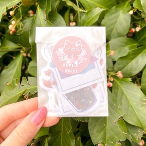 Puede incluir: Una bolsa de plástico transparente llena de pegatinas coloridas. Las pegatinas muestran un gato de dibujos animados, un diseño de galaxia y un marco de fotos con el texto "oopsie daisy".