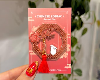 Könnte beinhalten: Eine roségoldene Emaille-Anstecknadel mit einem weißen Kaninchen, das unter einem rosa Kirschblütenbaum in einem sechseckigen Rahmen mit einem roten Quasten sitzt. Die Anstecknadel ist mit "Chinese Zodiac - Enamel Pin" und "CRAFTS.COM" beschriftet.