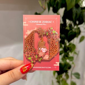 Może przedstawiać: Złota emaliowana przypinka przedstawiająca małpę wiszącą na gałęzi kwitnącej wiśni. Przypinka jest otoczona różowo-zielonym wzorem kwiatowym i ma czerwone tło z tekstem "CHINESE ZODIAC - Enamel Pin -" i "www.doozicrafts.com".
