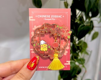May include: A Chinese Zodiac enamel pin with a yellow cartoon character, red flowers, and a gold frame. The pin has a yellow tassel and a red background with the text "CHINESE ZODIAC - Enamel Pin -."