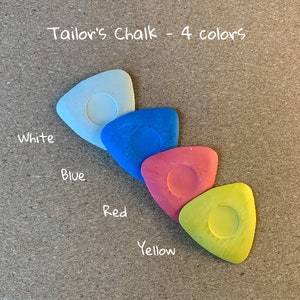 May include: Four tailor's chalk pieces in a triangular shape, arranged in a row from white to blue, red, and yellow. The text "Tailor's Chalk - 4 colors" is written above the chalk pieces.