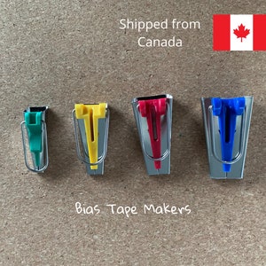 May include: Four bias tape makers in different colors: green, yellow, red, and blue. The bias tape makers are made of metal and have a small plastic piece at the top. The bias tape makers are arranged in a row on a brown surface.  Bias Tape Makers.