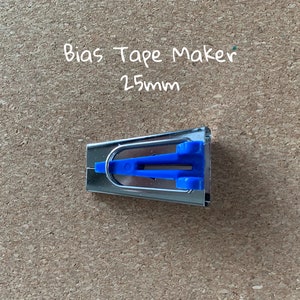 K&ouml;nnte beinhalten: Ein silberner Schr&auml;gbandmacher mit blauen Kunststoffgriffen. Der Text "Bias Tape Maker 25mm" ist in Wei&szlig; auf braunem Hintergrund geschrieben.
