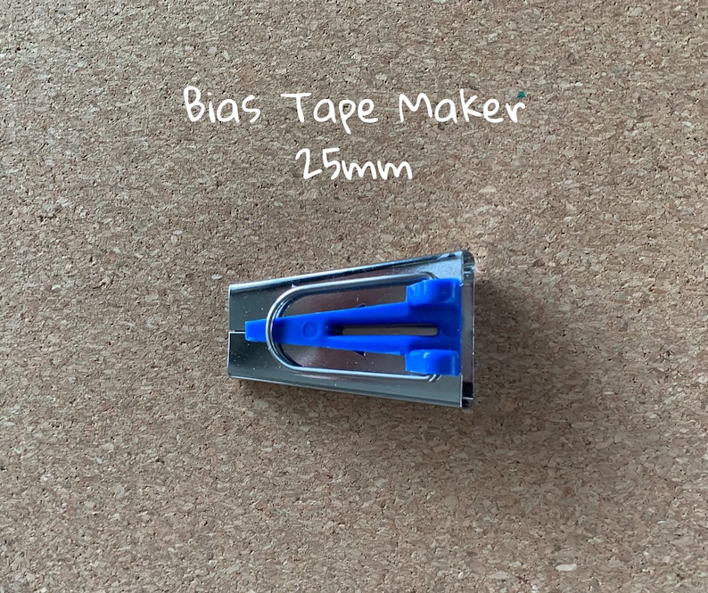 K&ouml;nnte beinhalten: Ein silberner Schr&auml;gbandmacher mit blauen Kunststoffgriffen. Der Text "Bias Tape Maker 25mm" ist in Wei&szlig; auf braunem Hintergrund geschrieben.