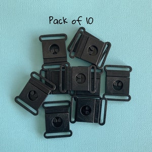 Pu&ograve; includere: Un pacco di dieci fibbie di plastica nera, ciascuna con un telaio rettangolare e un design circolare centrale. Le fibbie sono disposte su una superficie strutturata azzurra. Il testo "Pack of 10" &egrave; visibile.