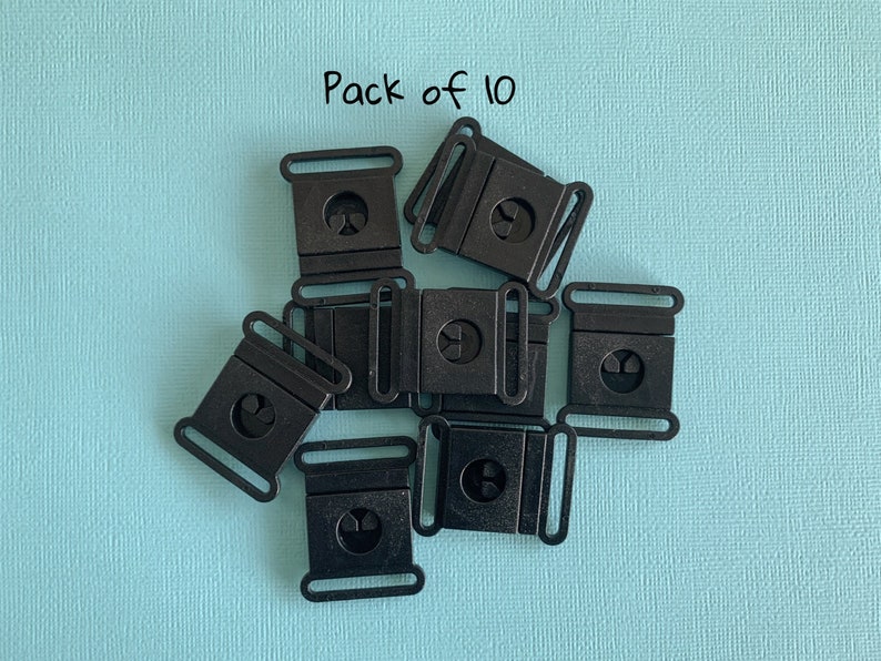 Pu&ograve; includere: Un pacco di dieci fibbie di plastica nera, ciascuna con un telaio rettangolare e un design circolare centrale. Le fibbie sono disposte su una superficie strutturata azzurra. Il testo "Pack of 10" &egrave; visibile.