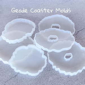 May include: Five clear silicone molds shaped like geode coasters. The molds are stacked on top of each other. The text "Geode Coaster Molds" is written in white on a gray background.