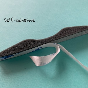 Peut inclure: Gros plan sur une bande de mousse noire avec un dos adh&eacute;sif blanc. Le texte "self-adhesive" est &eacute;crit au-dessus de la bande.
