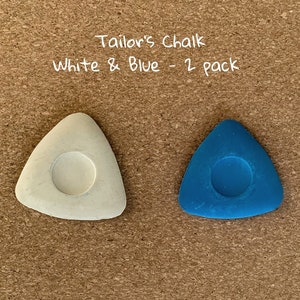 May include: Two tailor's chalk pieces, one white and one blue, in the shape of a triangle with a round hole in the center. "Tailor's Chalk White & Blue - 2 pack" is written above the chalk.