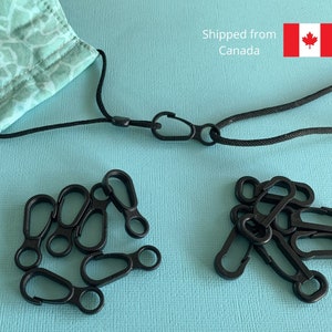 May include: A set of black plastic clips for attaching a face mask to a lanyard. The clips are in a pile on a light blue surface. The lanyard is black and has a metal ring on one end.