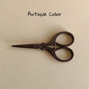 May include: Antique color small scissors with ornate details. The scissors have a brown finish and are closed.