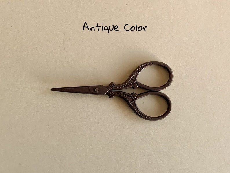 May include: Antique color small scissors with ornate details. The scissors have a brown finish and are closed.