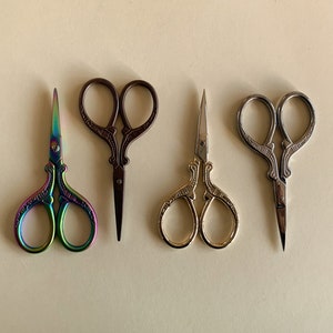May include: Four pairs of decorative scissors with ornate handles. The scissors are in different colors: rainbow, brown, gold, and silver.