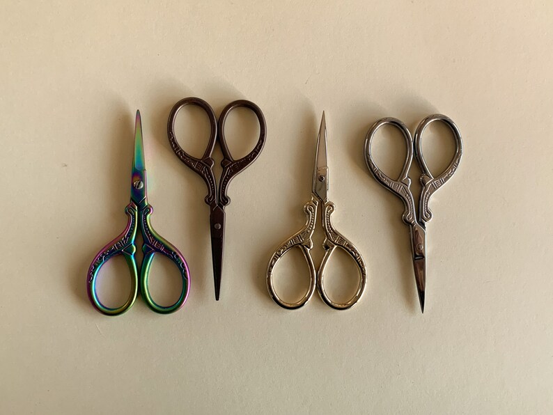 May include: Four pairs of decorative scissors with ornate handles. The scissors are in different colors: rainbow, brown, gold, and silver.
