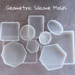 May include: A set of nine white silicone moulds in various geometric shapes, including circles, squares, octagons, and rectangles. The moulds are arranged on a grey textured surface. Geometric Silicone Moulds.