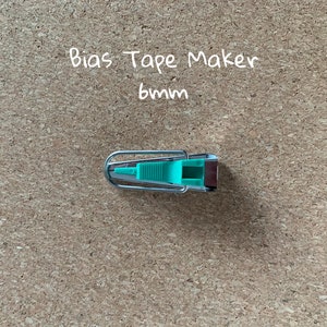 K&ouml;nnte beinhalten: Ein silberner Schr&auml;gbandmacher mit einem gr&uuml;nen Kunststoffgriff. Der Text "Bias Tape Maker 6mm" ist &uuml;ber dem Werkzeug geschrieben.