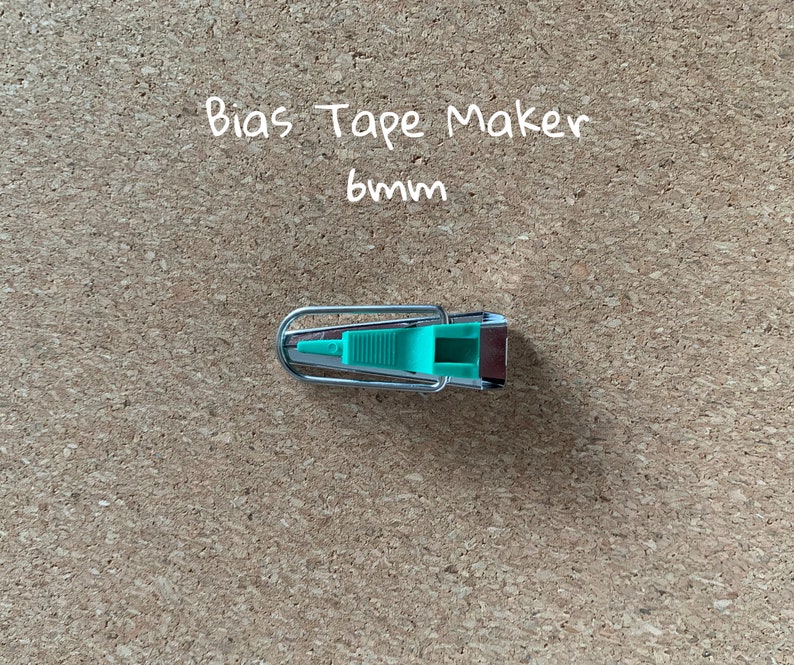 K&ouml;nnte beinhalten: Ein silberner Schr&auml;gbandmacher mit einem gr&uuml;nen Kunststoffgriff. Der Text "Bias Tape Maker 6mm" ist &uuml;ber dem Werkzeug geschrieben.